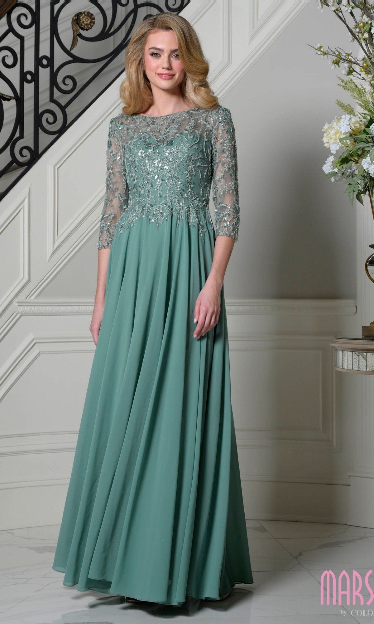 A woman stands indoors wearing the Long Formal Dress: Marsoni M340SL in sage, featuring a beaded-embroidered bodice, sheer three-quarter sleeves, and a graceful a-line chiffon skirt.